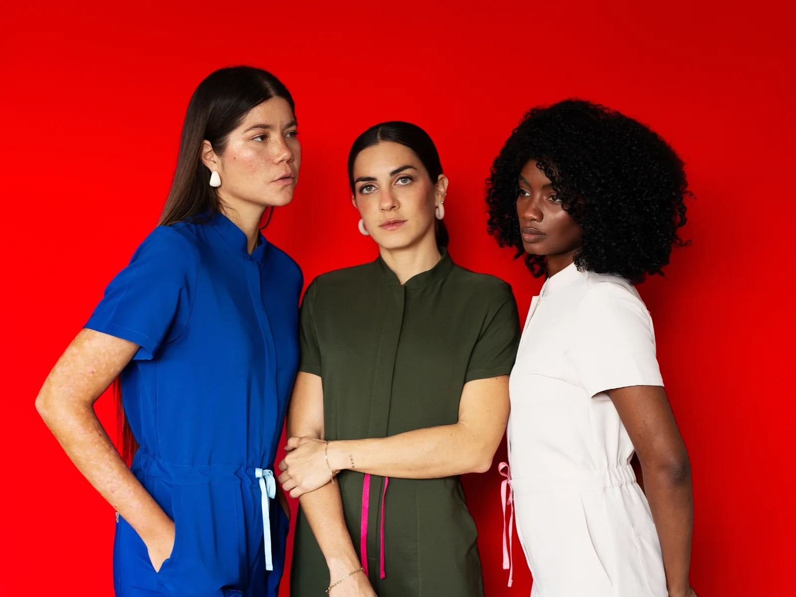 Tres mujeres con scrubs en azul, verde y blanco posando frente a un fondo rojo.